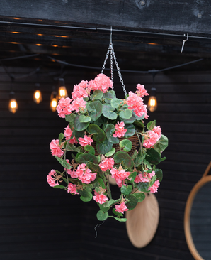 Le Bégonia artificiel rose en panier UV 70cm, avec des feuilles vertes luxuriantes et des fleurs roses, est suspendu à une chaîne. Des lumières rasantes et un miroir rond accentuent le fond en bois sombre.
