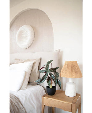 Une chambre confortable aux tons neutres comprend un lit avec des draps blancs, une table de chevet en bois avec l'Art Begonia 48 cm en pot décoratif noir et une lampe tressée, et une décoration murale ronde et texturée au-dessus du lit.