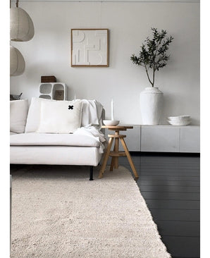 Un salon minimaliste avec un canapé de couleur claire, un tapis neutre, un buffet blanc, un olivier artificiel de 120 cm comme élément décoratif, une décoration murale abstraite et des tabourets en bois - le tout dans une douce ambiance méditerranéenne aux tons neutres.