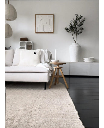 Un salon minimaliste avec un canapé de couleur claire, un tapis neutre, un buffet blanc, un olivier artificiel de 120 cm comme élément décoratif, une décoration murale abstraite et des tabourets en bois - le tout dans une douce ambiance méditerranéenne aux tons neutres.