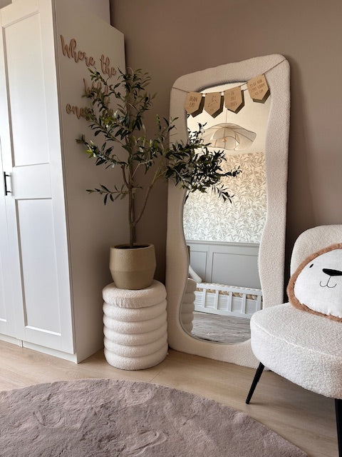 Une chambre de bébé confortable et moderne dans des tons neutres comprend un miroir de sol ondulé, un olivier artificiel de 120 cm pour une touche méditerranéenne, une chaise moelleuse avec un coussin en forme de tête d'ours et un berceau reflété dans le miroir - le décor parfait pour n'importe quelle pièce.