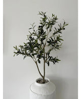 L'Olivier Artificiel 120 cm présente des feuilles vertes et des olives noires dans un pot blanc avec structure et galets, ce qui en fait un bel élément de décoration sur un mur uni gris clair.