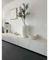 Une pièce minimaliste avec un buffet blanc, l'Olivier Artificiel 120 cm comme élément décoratif, des bols décoratifs blancs, une bougie et une décoration murale abstraite. Les parquets en bois sombre et les accents modernes créent une atmosphère méditerranéenne sereine.