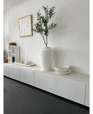 Une pièce minimaliste avec un buffet blanc, l'Olivier Artificiel 120 cm comme élément décoratif, des bols décoratifs blancs, une bougie et une décoration murale abstraite. Les parquets en bois sombre et les accents modernes créent une atmosphère méditerranéenne sereine.