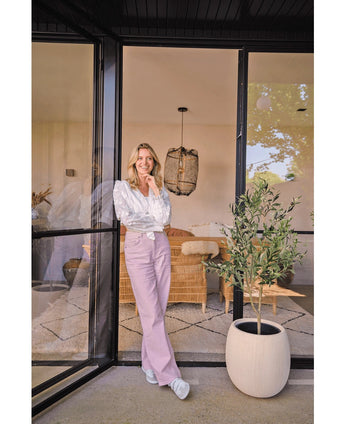 Une femme vêtue d'un chemisier blanc et d'un pantalon lavande sourit près d'une grande porte vitrée. L'Olivier Artificiel 120 cm apporte une ambiance méditerranéenne au salon cosy, avec un canapé en osier et une décoration lumineuse en fond.