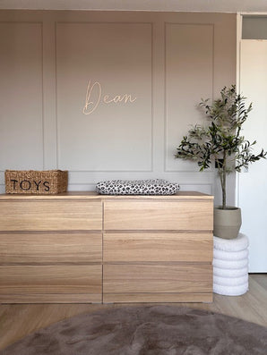 Une chambre d'enfant minimaliste contient une commode en bois avec un matelas à langer à imprimé léopard, un panier tressé étiqueté "Toys" comme décoration, l'Olivier Artificiel 120 cm pour le vert et "Dean" en cursive sur le mur beige au-dessus.