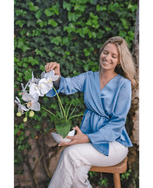 Une femme souriante vêtue d'un chemisier bleu clair et d'un pantalon blanc est assise sur un tabouret, tient l'orchidée artificielle 50 cm blanche dans un pot et touche doucement les fleurs. Elle est assise sur un fond de feuillage vert.