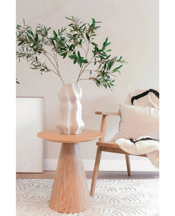 Un salon moderne avec une chaise en bois avec un coussin léger, une table d'appoint ronde et un vase sculptural blanc avec feuille d'olivier artificielle 90 cm UV, créant un espace minimal aux tons neutres doux et une subtile atmosphère méditerranéenne.