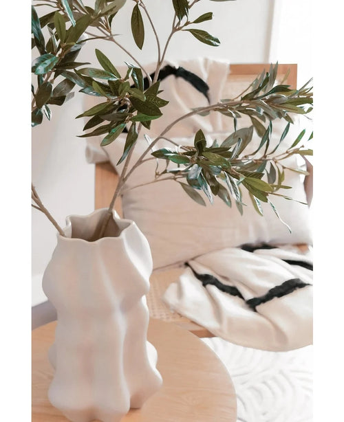 Un vase sculpté blanc contient une feuille d'olivier Art 90 cm UV avec des feuilles vertes sur une table en bois clair. Derrière elle, une chaise beige et deux coussins de couleur crème aux rayures noires éclatantes rehaussent l'atmosphère méditerranéenne.