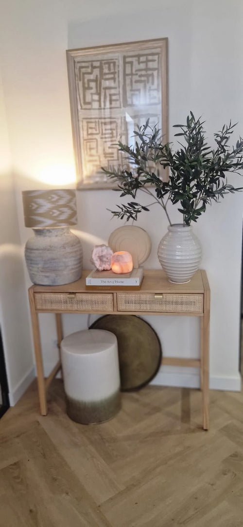 Une console en bois avec deux tiroirs contient une lampe, un grand vase blanc avec feuille d'olivier artificielle de 90 cm ignifuge, un plus petit vase, des livres et des pierres décoratives. Des accents méditerranéens et des œuvres d’art abstraites encadrées complètent le look ci-dessus. En bas se trouvent un tabouret et un plateau rond.