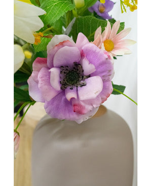 Un gros plan de la fleur d'anémone artificielle violette Real Touch de 52 cm dans un vase blanc mat, entourée de feuilles vertes et de fleurs aux couleurs pastel.
