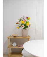 Un vase beige avec des fleurs artificielles d'anémone Real Touch violettes de 52 cm se dresse sur une étagère en bois clair à côté d'une petite horloge ronde et d'un plateau. Au premier plan se trouve une table ronde blanche contre un mur et un rideau blancs.