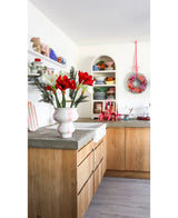Une cuisine lumineuse avec des armoires en bois, des étagères blanches ouvertes avec des assiettes et des casseroles, une couronne festive au mur et une fleur artificielle Amaryllis Real Touch rouge 36 cm sur le plan de travail en béton.