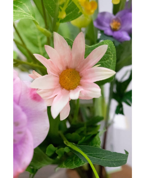 Un gros plan de la fleur artificielle Aster de 60 cm, aux pétales roses avec un centre jaune, entourés de feuilles vertes et d'autres fleurs, dont une violette, en arrière-plan.