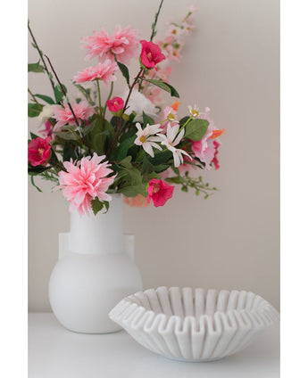 Une fleur artificielle Dahlia de 68 cm rose dans un vase blanc se trouve à côté d'un bol en céramique blanche avec des volants sur une surface blanche, sur un fond uni clair.
