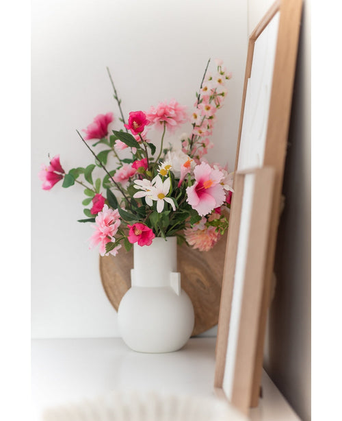 Une marguerite artificielle de 60 cm blanc/rose se dresse dans un vase blanc sur une surface blanche à côté d'une œuvre d'art dans un cadre en bois, avec un plateau rond en bois visible en arrière-plan.