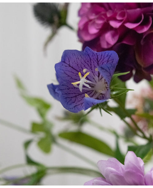 Un gros plan de la fleur artificielle Harebell 62 cm violette, une délicate fleur artificielle violette avec des nervures profondes et des étamines jaunes, entre des feuilles vertes et des fleurs rose pâle et violettes en arrière-plan.