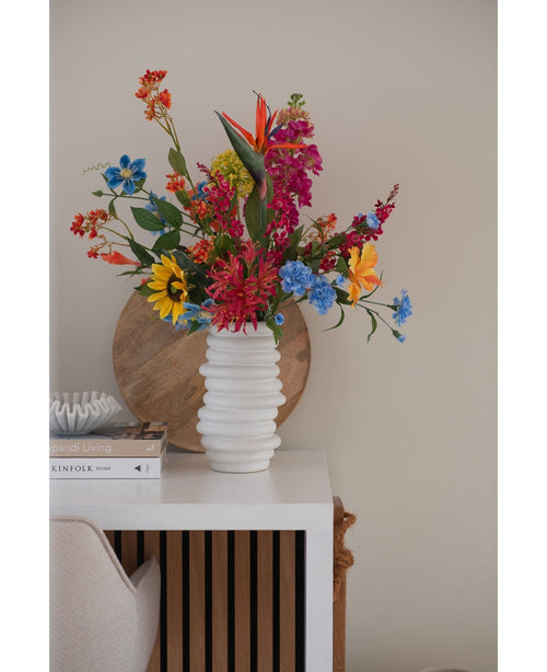 Un vase blanc texturé contient un bouquet rose d'herbe à chat artificielle de 75 cm sur une table blanche à côté de livres empilés et de décorations, avec un plateau rond en bois appuyé contre le mur beige en arrière-plan.