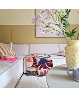 Un salon cosy avec un canapé beige clair, un plaid fleuri, une bougie rose, un coussin rayé et une fleur artificielle en dentelle de 76 cm blanche dans un vase jaune sur la table ronde. L'art abstrait décore le mur derrière lui.