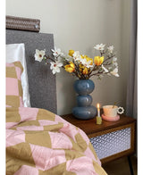 Une table de chevet avec un vase bleu contenant la Fleur Artificielle Magnolia 50 cm jaune/orange, une tasse à thé rose sur une soucoupe en forme de marguerite et une bougie allumée se dresse à côté d'un lit recouvert d'une couverture rose et marron aux motifs géométriques.