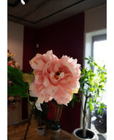 Un gros plan de la fleur artificielle pivoine 61 cm rose tendre, exposée à l'intérieur avec des feuilles vertes et des plantes en pot en arrière-plan, baignée par la lumière naturelle des fenêtres.