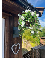 Le Bégonia Artificiel Blanc en Panier UV 70cm est accroché sur un mur en bois, avec un cœur blanc décoratif en dessous. De la verdure, une terrasse en bois et un ciel bleu partiellement nuageux complètent le décor.