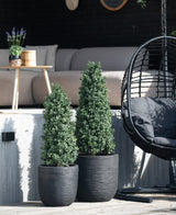 Deux plantes artificielles de buis pyramide UV de 120 cm sont placées sur une terrasse en bois devant un coin salon extérieur avec un canapé lumineux, une table ronde avec de la lavande et un fauteuil suspendu noir.
