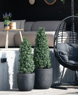 Deux plantes artificielles de buis pyramide UV de 120 cm sont placées sur une terrasse en bois devant un coin salon extérieur avec un canapé lumineux, une table ronde avec de la lavande et un fauteuil suspendu noir.