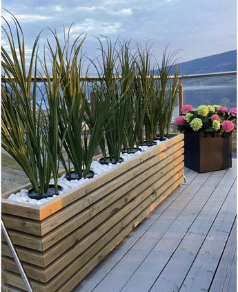Une jardinière en bois avec du gazon artificiel vert UV 90 cm et des pierres blanches se dresse sur une terrasse en bois à côté d'une jardinière noire avec des hortensias colorés, avec des balustrades en verre et une vue sur les montagnes en arrière-plan.