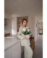 Une femme aux cheveux blonds bouclés, vêtue d'une tenue vert clair, se tient dans une cuisine moderne avec dans ses mains une plante d'hortensia artificiel blanc UV de 35 cm – avec des fleurs blanc crème – et sourit à la caméra.
