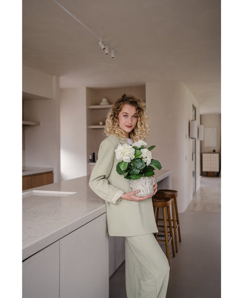 Une femme aux cheveux blonds bouclés, vêtue d'une tenue vert clair, se tient dans une cuisine moderne avec dans ses mains une plante d'hortensia artificiel blanc UV de 35 cm – avec des fleurs blanc crème – et sourit à la caméra.