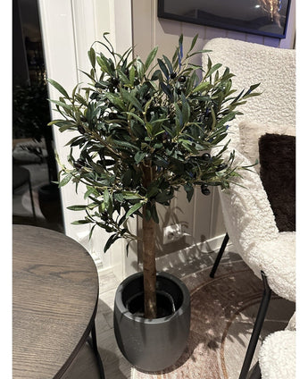 L'Olivier artificiel UV 85 cm, une plante artificielle compacte et facile d'entretien aux feuilles vert foncé et aux olives noires, se dresse en intérieur à côté d'une table ronde en bois et d'un fauteuil à la texture blanche.