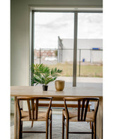 Une table en bois avec deux chaises, un salon de palmiers artificiels UV 60 cm dans un pot et un vase doré sont placés près d'une grande fenêtre donnant sur une zone industrielle avec des bâtiments et une clôture.