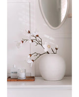 Une branche de magnolia artificielle Real Touch White 98 cm se dresse dans un vase rond blanc sur une surface blanche à côté d'une bougie en verre allumée et d'une petite bouteille sur un plateau en bois, dans une pièce lumineuse et minimaliste avec un miroir rond au-dessus.