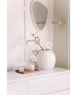 Une branche de magnolia artificielle blanche Real Touch White 98 cm se dresse dans un vase sur un buffet blanc à côté d'un petit verre, d'un flacon de parfum et d'un plateau en bois. A l'étage, un miroir de forme irrégulière est accroché au mur de couleur claire.