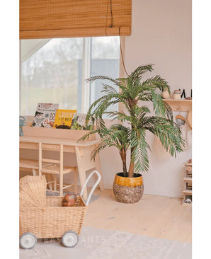 Dans une pièce cosy et lumineuse, les palmiers artificiels Cycas Classic 135 cm sont placés dans une jardinière élégante, un chariot à jouets en osier, des meubles en bois et des livres pour enfants près d'une fenêtre avec un store enrouleur en bambou.