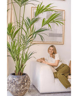Une femme aux cheveux blonds, vêtue d'un haut crème et d'un pantalon vert olive, est assise sur un canapé blanc, souriante et tenant une tasse. Le palmier artificiel Kentia 220 cm rehausse l'atmosphère tropicale de la pièce lumineuse et cosy avec une décoration murale abstraite.