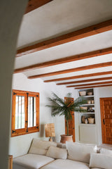 Un salon confortable avec des murs blancs, des poutres en bois, un canapé sectionnel blanc, le palmier artificiel Phoenix De Luxe 210 cm, une fenêtre et une porte en bois, des étagères intégrées avec des paniers et des poteries et de la lumière naturelle entrant par la fenêtre.