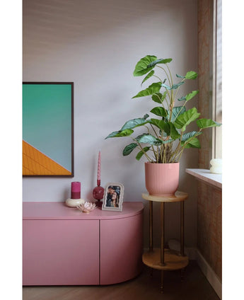 Un coin salon moderne avec un buffet rose, des bougies, une décoration murale géométrique, une photo encadrée et la plante artificielle Feuille de pommier de 90 cm - une grande plante à feuillage dans un pot rose sur un petit support en bois près de la fenêtre.