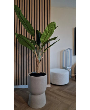 Le bananier artificiel de 150 cm, avec ses feuilles vertes luxuriantes, se dresse sur un plancher en bois près d'un mur nervuré avec des panneaux de bois. A proximité se trouve un pouf rond blanc et un ventilateur sans pales en arrière-plan.