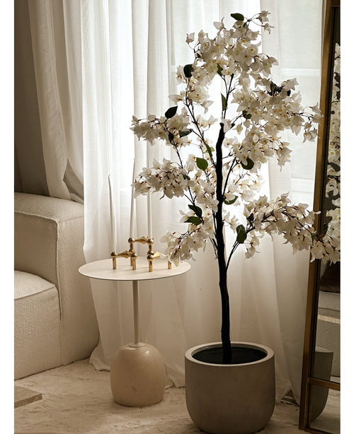 Une plante de bougainvilliers artificiels mesurant 120 cm se trouve près d'un miroir bordé d'or, à côté d'une petite table d'appoint ronde blanche avec des chandeliers dorés, de hautes bougies blanches, des rideaux blancs transparents et un canapé de couleur crème dans une pièce lumineuse.