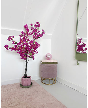 Un coin de pièce moderne et minimaliste avec une plante violette artificielle de bougainvilliers de 125 cm dans un pot, un repose-pieds rond rose avec des pieds dorés, des livres empilés, une boule de cristal rose, un miroir mural et un tapis rose tendre sur un sol clair.
