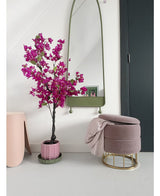Un intérieur moderne avec le bougainvillier artificiel 125 cm violet dans un pot décoratif, un grand miroir avec un cadre vert, un repose-pieds rose avec base dorée et couverture rose, et une porte sombre contre un mur blanc éclatant.