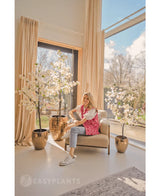 Une femme vêtue d'un cardigan à motifs roses et d'un jean est assise sur un fauteuil beige près de grandes fenêtres avec des rideaux de couleur crème, entourée de trois bougainvilliers artificiels blancs de 180 cm dans des jardinières dorées brillantes. La lumière du soleil remplit la pièce moderne et aérée.