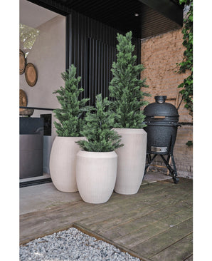 Trois jardinières hautes et blanches avec une plante de conifère artificiel UV de 90 cm se trouvent sur une terrasse en bois à côté d'un bâtiment moderne. Un gril en céramique noire se dresse à proximité, contre un mur de pierre bordé de vignes.