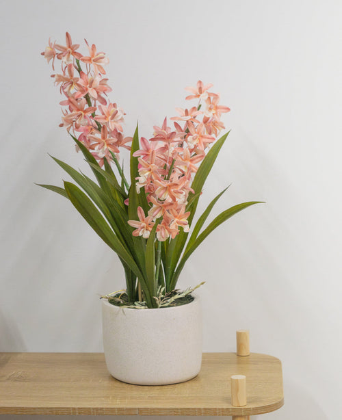 La plante artificielle Cymbidium rose de 58 cm, une plante verte avec de longues feuilles et de longues tiges avec des fleurs rose clair dans un pot en céramique blanche, se dresse sur une table en bois clair contre un mur blanc uni.