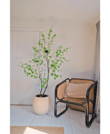 Dans un coin de pièce minimaliste, la plante artificielle Enkianthus 210 cm, une plante feuillue verte en pot, se dresse à côté d'une chaise en rotin avec un cadre noir sur un plancher en bois blanc, avec des murs neutres et un rideau beige clair.