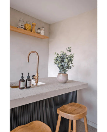 Un comptoir moderne comprend un robinet en laiton, des distributeurs de savon, une plante en pot d'eucalyptus artificiel de 61 cm et deux tabourets en bois. Au-dessus du comptoir, des étagères sur un mur clair accueillent des bocaux et des bouteilles.