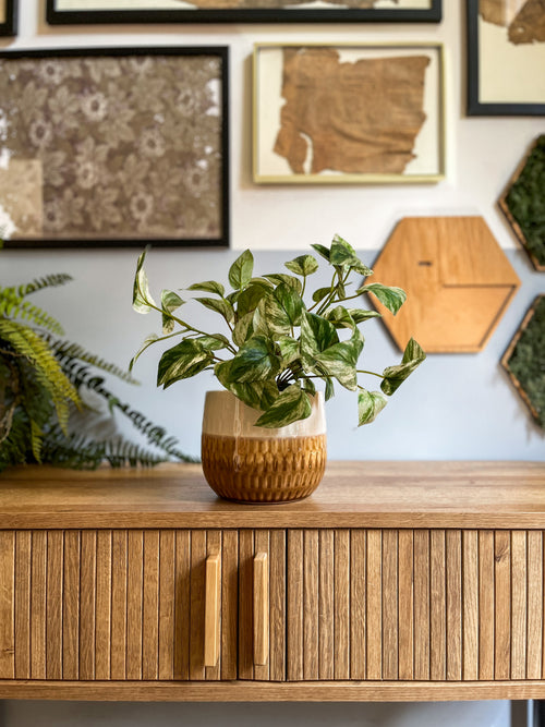 La Plante Artificielle Vigne Dorée en Pot 25 cm se dresse sur une armoire en bois avec des rainures verticales, avec un art encadré et une décoration murale hexagonale sur le mur de couleur claire derrière elle.