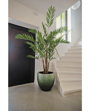 La plante artificielle Hawaii Parlour 180 cm, un grand palmier artificiel d'intérieur aux feuilles vertes luxuriantes, se dresse sur un sol gris dans un hall moderne, près d'un escalier blanc et d'une porte noire.